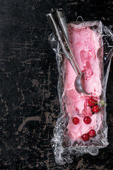 Homemade cherry sorbet in vintage iron container with polyethylene film, fresh cherry berries and metal spoon for ice cream over old black wooden textured background. Top view with copy space