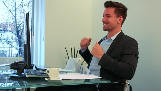 A young, handsome man works on a computer in an office, then throws papers around and starts to celebrate