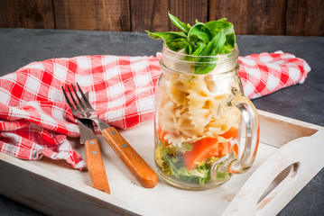 Italian salad with pasta, basil, cheese, tomato and lettuce. In mason jar, on a tray with a towel, knife and fork; on the gray stone table, copy space