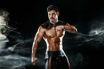 Portrait of handsome muscular gladiator with sword. Studio shot. Black background. - obrazy, fototapety, plakaty