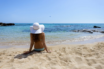 Fototapeta na wymiar Young woman sitting on the beach. Portrait of a girl in white hat relaxing at tropicon beach. Beautiful Summer sea side beach with turquoise water. View from behind 