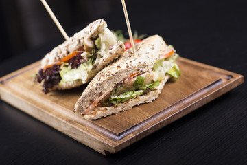 Tasty grilled chicken sandwiches with a wooden stick stabbed trough them on a wooden plate. Meat sandwiches with lettuce, tomatoes and french fries on the side.