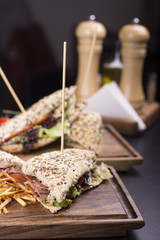 Tasty grilled sandwiches with a wooden stick stabbed trough them on a wooden plate. Meat sandwiches with lettuce, tomatoes and french fries on the side. Napkins and olive oil on the background.