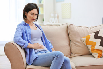 Beautiful young woman suffering from abdominal pain while sitting on sofa at home