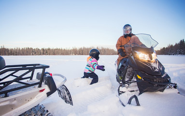 A four year old girl in winter coat and ski pants wearing a helmet walking through deep snow toward her father on a snowmobile parked in a white snow covered field 
