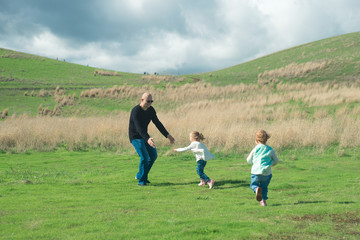 Smiling father playing and running with his two laughing daughters in the park on green grass. Beautiful view with stormy clouds over their heads.