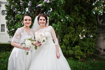 Bride with bridesmaid looking at camera near lilac tree.