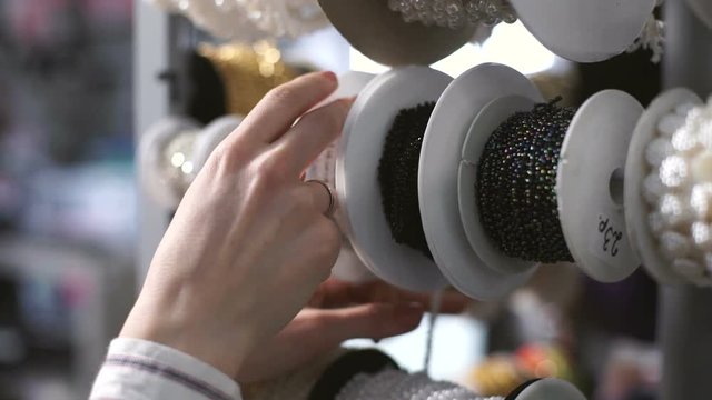 Tailor selects accessories with windows for decoration of clothes. On a shelf hanging white spinning reels with different shiny ribbons. Hands stop rack and seamstress begins to pick something that is