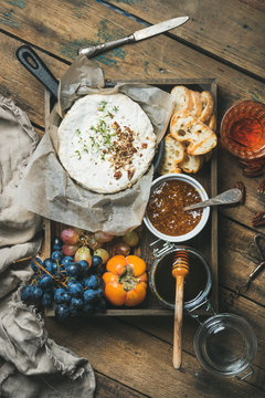 Cheese, fruit and wine set. Camembert in small pan, grapes, persimmon, fig jam, honey, baguette slices and glass of rose wine over rustic wooden background, top view, vertical composition