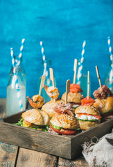 Home party food concept. Homemade burgers in wooden tray and lemonade in glass bottles with straws on rustic table, bright blue wall at backgound, selective focus, copy space