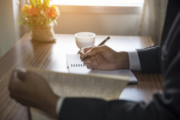 Smart man reading and writing something on note book .He sitting at a wooden desk with a coffee cup...