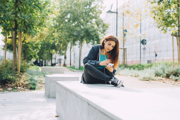 Young handsome caucasian reddish hair woman sitting on a small wall, listening music with headphones and smartphone handhold - music, relax, technology concept