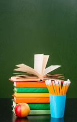 Stack of books with apple and pencils near empty green chalkboard