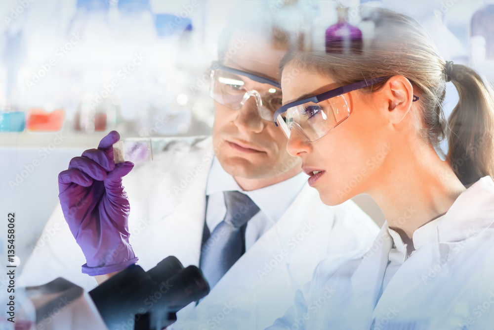 Poster Attractive young scientist and her post doctoral supervisor looking at the microscope slide in the forensic laboratory.