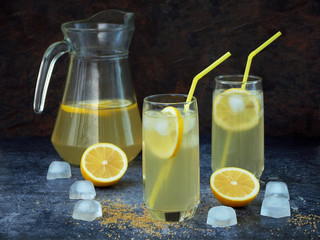 Two glasses and carafe of cold homemade lemonade with lemon slices, ice cubes, brown sugar, yellow straws on dark background.