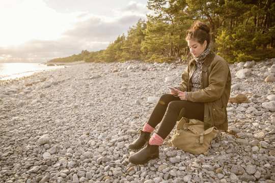 Woman sitting on rock with bag while using mobile phone at beach