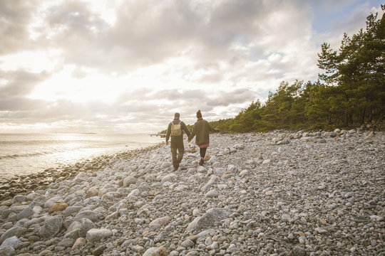 Rear view of couple walking on rocky shore against sky during sunset