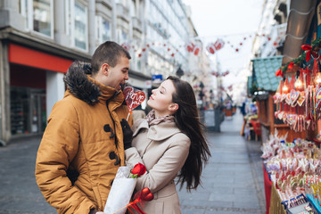 Beautiful young couple in love enjoying together at city street, kissing and holding together a lollipop in heart shape. 