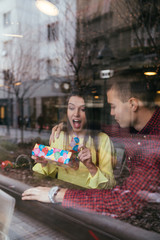 Beautiful young couple in love enjoying together at cafeteria, and man is giving a gift as surprise to his girlfriend. 