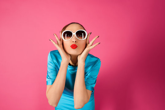 portrait of shocked and surprized young beautiful woman in blue silk dress and white sunglasses ot the pink background