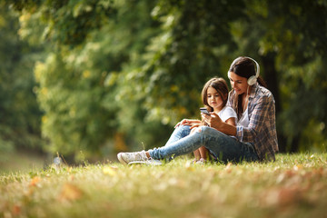 Mother and daughter sitting on grass and listening to music on smart phone.