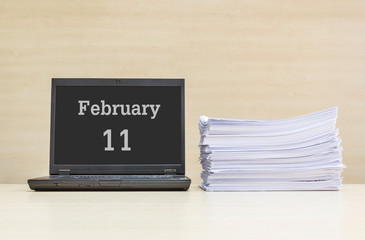 Closeup computer laptop with february 11 word on the center of screen in calendar concept and pile of work paper on wood desk and wood wall in work room textured background with copy space