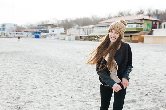 Picture of young woman walking in beach with cute smile and long hair. She is wearing in knit cap and dark jacket.
