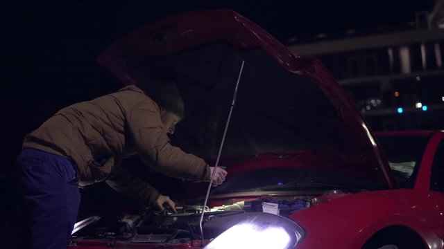 Man looking at engine of broken down car with flashlight at night. Headlights turned on