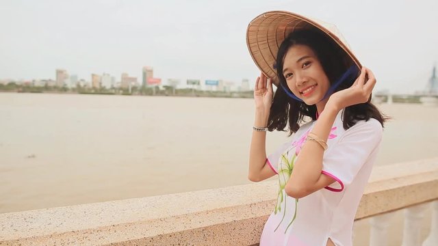Vietnam girl in the national costume and dress Ao Dai posing on the waterfront of the city of Da Nang. On the back of the cap Non La.