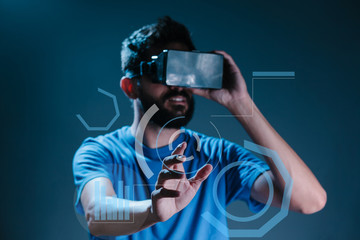 Obraz na płótnie Canvas Young bearded man in VR-headset over dark blue background is touching a virtual interactive screen
