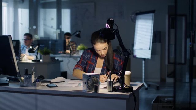 Late at Night Female Industrial Engineer Works on Drafts and Schemes Sitting at Her Desk. Her Colleagues Work in Background. Blue City Light Seeps Through Windows. Shot on RED Cinema Camera 4K (UHD).