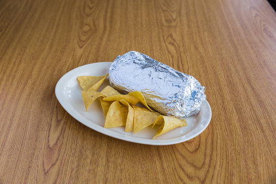 Foil Wrapped Burrito And Chips On White Plate On Wooden Table