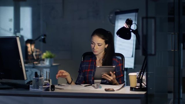 In Late Night Office Female Design Engineer Works on Drafts, She Consults Tablet Computer. Her Table is Lit by Lamp. Colleagues Work in the Background.  Shot on RED Cinema Camera 4K (UHD). 