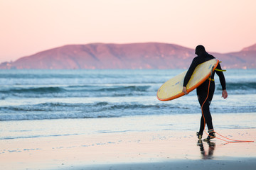 Surfer with surfboard and wetsuit walks towards the water at sunrise in Pismo Beach