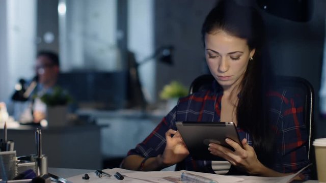 In Late Night Office Female Design Engineer Works on Her Drafts, She Consults Tablet Computer. Her Table is Lit by Lamp. Colleagues Work in the Background.   Shot on RED Cinema Camera 4K (UHD). 