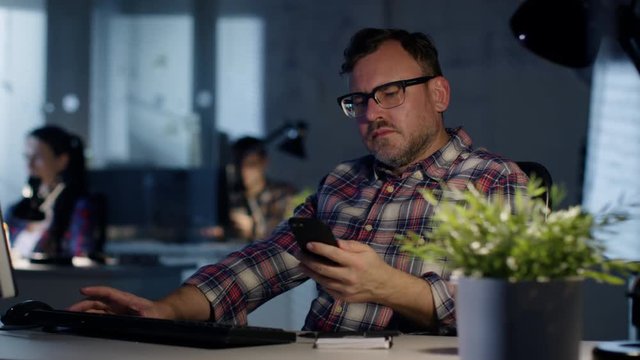Late Night at the Office. Man Sits at His Desk, Consults Smartphone, Writes Down a Note. In Background His Colleagues Work.  Shot on RED Cinema Camera 4K (UHD). 