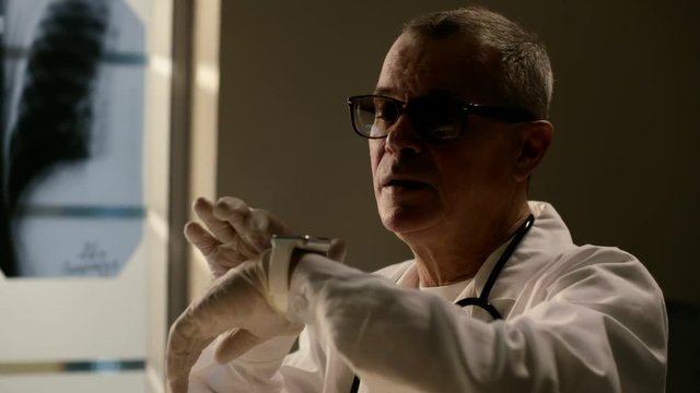 Ederly caucasian doctor in medical gown or robe with stethoscope using smart watch at modern office. Evening time. Shot was made for adding CG hologram.