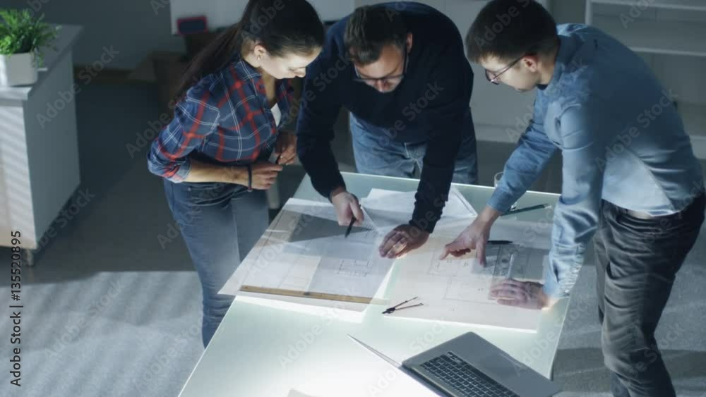 Poster late at night team of design engineers work with blueprints and documents on illuminated table. shot