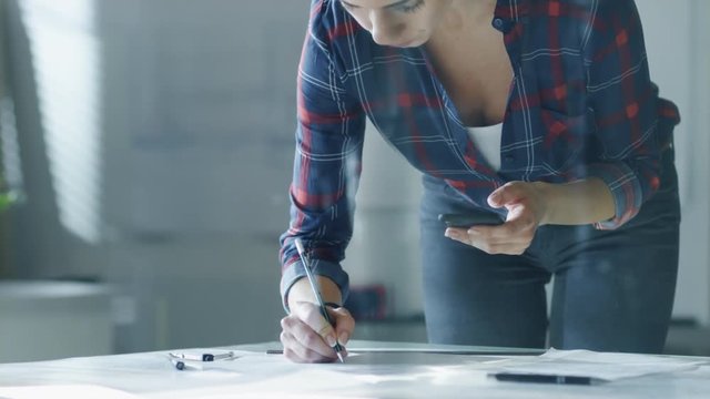Close-up of a Female Design Engineer Works on Documents in a Conference Room, Last Minute Check-up, Uses Her Smartphone. In the Background Whiteboard with Schemes on it, Various Blueprints on Walls.  