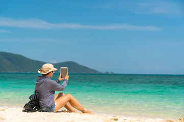hiker girl in a hat with a backpack with glasses sitting on the sand with a tablet in hands. making the photo on his tablet.
