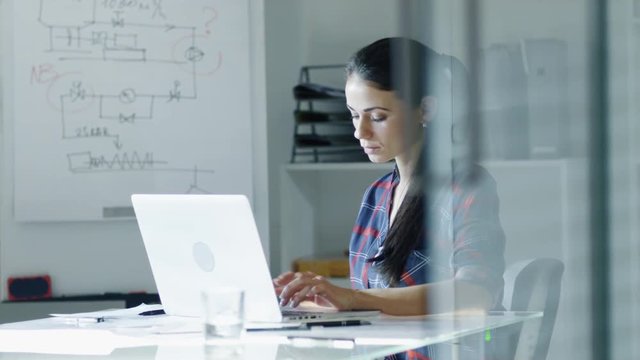 Female Design Engineer Sits at the Glass Table in Her Office, Types on a Laptop, Blueprints Laying on Her Desk