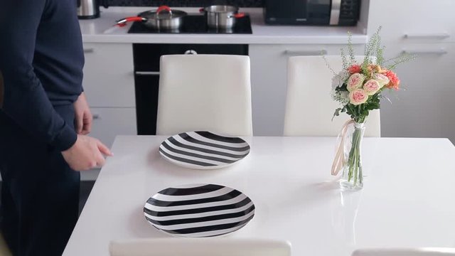 Man in the kitchen yourself serves white table for two persons. On the surface, on which stands a little delicate bouquet of flowers in a glass vase, Male puts a stylish striped plates and forks to