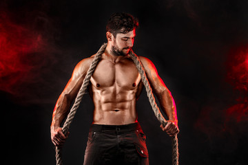 Muscular man holding rope and posing on the black background with red smoke