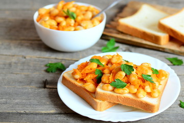 Baked white beans with vegetables on bread and on plate. Baked white beans in a bowl, bread slices on wooden background with copy space for text. Healthy vegetarian food
