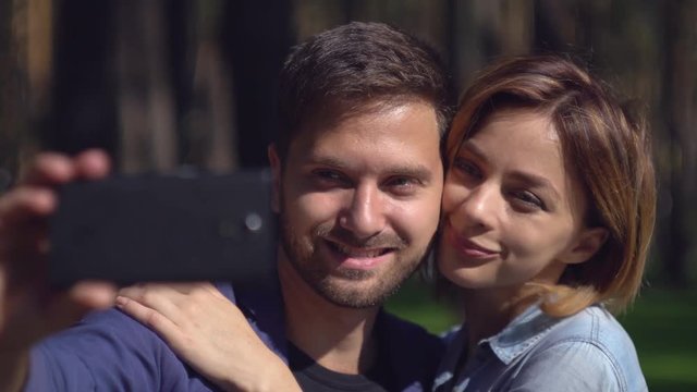 couple take selfie photo in park. Happy family have fun photographed with smartphone. Caucasian young man holding mobile phone. Smiling girl kissing boyfriend.