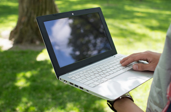 Man holding a laptop in one hand and prints it in the park. Sunny summer weather. Communication. Activities in the park. Freelancer.