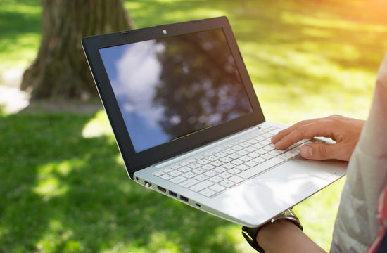 Man holding a laptop in one hand and prints it in the park. Sunny summer weather. Communication. Activities in the park. Freelancer.