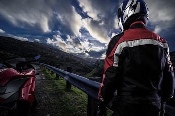 Fototapeta premium Male motorcyclist wearing protective leather racing suit with a red bike or motorcycle on an open road. The vehicle is cropped to become generic non branded. The image depicts travel and adventure.