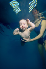 Obraz na płótnie Canvas Happy baby swims underwater in the pool on a blue background, while the mother supports him with his hands. Portrait. Shooting under water. Bottom view. Vertical orientation