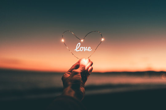 Simbolo del cuore illuminato con scritta love per San Valentino tenuto in mano di fronte al tramonto sul mare.